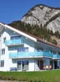 A multi-story apartment building with balconies, surrounded by a grassy area and with mountains in the background.