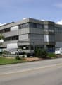Commercial building, grey facade, large windows, cars parked in front, greenery around