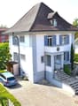 2 story house, white exterior, brown roof, blue windows, grey door, blue garage door, paved driveway, garden, lawn, hedge, mailbox, stairs, blue car parked