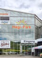 A large commercial building with a glass facade, featuring various retail and service establishments such as a sports store, an outlet store, a bistro, and a dairy and meat shop. The building is called 'Sport-Fabrik' and has a 'Hagi-Park' sign, indicating