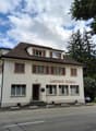 white house, brown roof, large windows, two cars parked, sign reads 'Gasthof Rossli', mountain in the background