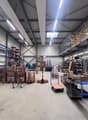 Spacious warehouse with high ceilings, multiple shelves and storage racks, various boxes and materials, trolleys and pallets, stairs, and metal beams.