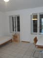 small room with single bed, wooden table, desk, wooden chair, lamp on table, white walls, windows, white tile floor