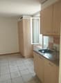 The image shows a kitchen area with beige cabinets, a sink, and a refrigerator. The floor is tiled, and there is a window visible. The kitchen appears to be part of a larger living space.