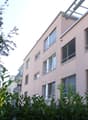 Multiple story building, pink paint, multiple windows with blinds, balcony, greenery