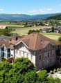 The image shows a scenic rural town with a mix of residential and commercial buildings. The buildings have red tile roofs and are surrounded by lush greenery, including trees and fields. There are several multi-story apartment buildings with balconies vis