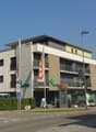 multi-story building with ground floor, pharmacy on ground floor, cars parked in front, greenery, streetlights, bicycles, bicycles