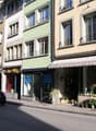 A street with buildings on both sides, a car driving, and a van parked on the side. The buildings have glass windows and balconies. The sidewalk has potted plants and a shop with a white awning. The shop has glass windows with potted plants and flowers di