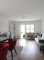 Open-concept living and dining area with hardwood floors, white walls, large windows, glass coffee table, red dining chairs, gray sofa, and hanging pendant lights. The room also features a dishwasher and various potted plants.