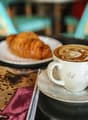 A cup of coffee with a latte art design, served with a croissant on a plate. The setting includes a newspaper and other items on the table, suggesting a cozy cafe environment.