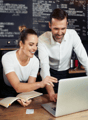 The image shows a couple working together at a cafe or restaurant. They are sitting at a counter, with a laptop and a book open in front of them. The background includes a chalkboard menu and other cafe equipment, suggesting this is a commercial food serv