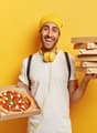 Man holding pizza boxes, smiling, wearing yellow headphones and a yellow hat