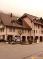 A row of multi-story residential buildings with red tile roofs, located in a mountainous area with fog in the background. The buildings have a traditional European architectural style with arched entrances and windows. There are cars parked on the street