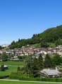 The image shows a picturesque mountain village nestled in a lush, green valley. The village is surrounded by rolling hills and dense forests, with a clear blue sky overhead. The buildings appear to be a mix of traditional and modern architecture, with red