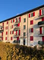 4 story apartment building, beige, red shutters, multiple balconies, hedges in front