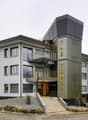 Contemporary building with multiple floors, modern facade, staircases leading to the entrance, car parked in front, and a surrounding landscape.
