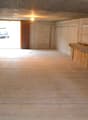 This image shows the interior of a garage or storage space. The space has concrete walls and a wooden floor. There is a window at the end of the room, allowing natural light to enter. The room appears to be empty and unfinished, with a simple light fixtur