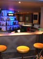 Modern bar with a sleek, geometric design. The bar counter is made of wood and has a granite-like surface. The back bar is well-stocked with various liquor bottles and glassware. There are yellow bar stools in front of the counter, and the lighting create