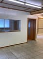 The image shows a large open office space with a tiled floor, white walls, and wooden beams on the ceiling. There is a window with blinds and a doorway leading to another room. The space appears to be a commercial or office setting.