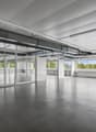 Large open office space with high ceilings, concrete floors, and floor-to-ceiling windows providing natural light and views of the surrounding greenery. The space has a modern, minimalist design with exposed ceiling beams and ductwork.