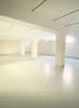 Large open space with white walls, concrete floor, and several windows providing natural light. The room appears to be an empty, unfinished space, possibly a loft or studio apartment.
