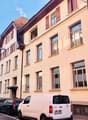 Multi-story building with beige walls, red shutters, many windows, ground floor and upper floors with balconies, white van parked in front.