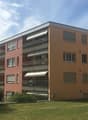5 story apartment building, beige and red facade, multiple balconies, fenced garden in front