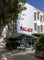 2-story building, white exterior, large glass windows, sign 'PALACE', plants, tree, sidewalk, cars