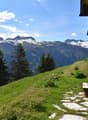 A wooden chalet situated on a grassy hillside with a stunning view of snow-capped mountains in the background. The chalet has wooden shutters and is surrounded by pine trees.