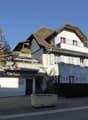 Multi-story residential building, white walls, brown roof, multiple balconies, ground floor entrance, staircase, hedge, large tree, small bench