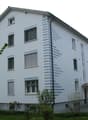 3-story white building, windows with shutters, chimney, entry area with canopy, greenery and plants surrounding the building