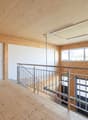 The image shows a large open space with a wooden ceiling and walls. There is a metal railing along the second floor balcony, and the floor is made of wood planks. The room has large windows that provide natural light. The overall design appears to be mode