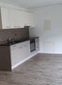 Open-concept studio apartment with white walls, hardwood-style flooring, and a modern kitchen with white cabinets, a black countertop, and a built-in dishwasher.