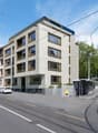 Multistory residential building, modern design, several windows, elevated ground floor, street tram tracks, bus stop, trees, parked cars