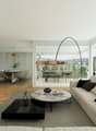 Spacious open-plan living area with large windows, hardwood floors, modern furniture including a sofa, coffee table, and dining table with chairs. The room has a balcony or terrace overlooking the city.