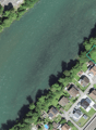 This image shows an aerial view of a residential area along a river or stream. The area is surrounded by trees and greenery, with a mix of single-story and multi-story homes. Some of the homes appear to have balconies or terraces visible. The river or str