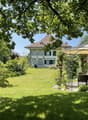 Large house with brown roof, white walls, multiple windows, surrounded by a well-maintained garden with a gazebo, table, and chairs.