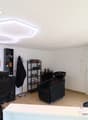 Spacious hair salon with modern decor, including black salon chairs, shelves with hair products, and a large circular mirror. The room has a bright, open layout with recessed lighting fixtures on the ceiling.