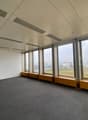 Large open office space with floor-to-ceiling windows providing a view of the surrounding cityscape. The room has a gray carpeted floor and a suspended ceiling with recessed lighting. There are built-in benches along the windows.
