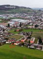 Aerial view of a neighborhood with multiple residential buildings and houses, a central green space, and surrounding fields and mountains.