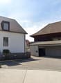 A multi-story residential building with a garage and parking spaces in the foreground. The building has a mix of modern and traditional architectural styles, with a combination of flat and sloped roofs. There are several windows and balconies visible on t