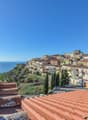 Picturesque coastal town with colorful buildings, red tile roofs, and a church steeple overlooking the sea. The town is situated on a hillside with lush greenery surrounding it.