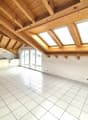 Spacious open-plan living area with vaulted wooden ceiling, large windows, and sliding glass doors leading to a balcony or terrace. The kitchen is located in the corner with white cabinets and tiled flooring throughout the space.