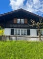 2 story chalet house with brown roof, white walls, wooden shutters, and wooden fence in front, surrounded by grass