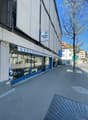 Commercial building with a shopfront, blue awning, and a sign reading 'Messer Wyss'. The building has a modern design with a white exterior and blue accents. The shopfront features large glass windows displaying items inside. There is a parking sign and a