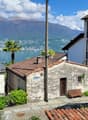 The image shows a picturesque view of a small town nestled along the shores of a lake, with mountains in the background. The buildings have traditional tile roofs and stone walls, and there are palm trees visible. The scene has a charming, quaint atmosphe