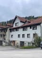 Large building with multiple floors, tiled brown roof, white walls, several windows, located in a scenic setting with green fields and trees