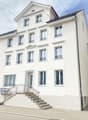 White building with multiple windows, stairs leading to the entrance, car parked in front, small garden with plants