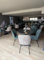 Modern indoor dining area with round tables and chairs, wooden floor, round lamps on tables, decorative plants, bar counter with chairs, and artwork on the wall.