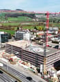 Aerial view of a construction site with a large red crane, surrounded by various buildings and a road with cars.